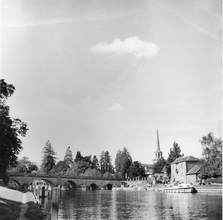 A black and white photograph of an old bridge across the river Thames at Wallingford, Oxfordshire. The spires of a town are in the background and boats are moored on the river banks